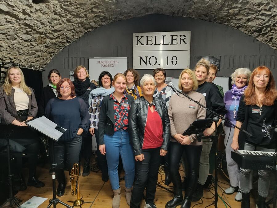 Die Frauen mit Leiterin Helga Forster in der Mitte stehen vor einem Schild mit der Aufschrift Keller NO. 10. Im Vordergrund sind Musikinstrumente zu sehen.