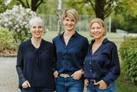 Man sieht 3 l&auml;chelnde Frauen. Sie sehen in die Kamera. Hinter ihnen sieht man B&auml;ume, B&uuml;sche und Wiese.