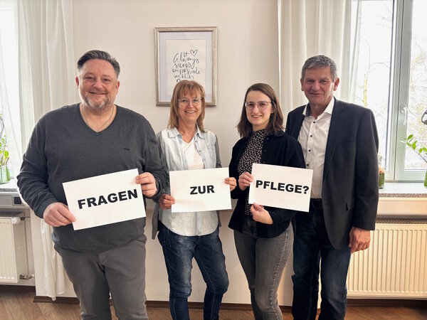 Vier Menschen stehen vor einer Fensterfront und halten Papier in den H&auml;nden. Der Text auf dem Papier hei&szlig;t "Fragen zur Pflege?"