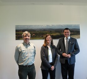 Zwei M&auml;nner und eine Frau stehen vor einem Bild und blicken in die Kamera
