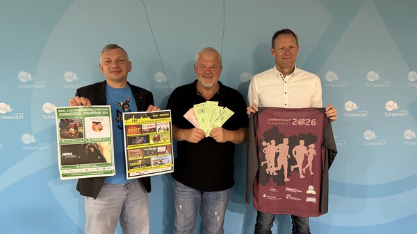 Vor einem blauen Hintergrund stehen drei M&auml;nner. Sie halten Plakate, Eintritts-Karten und ein Lauf-Shirt in den H&auml;nden.