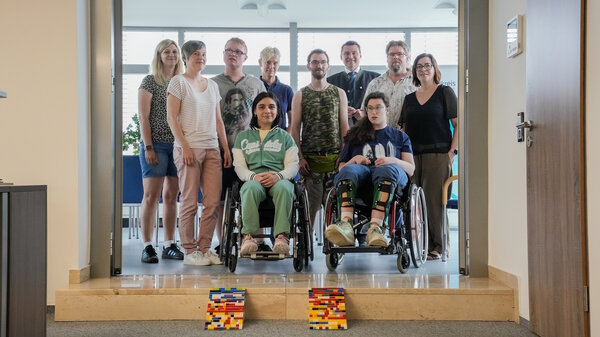 Das Bild zeigt M&auml;nner und Frauen auf einer Stufe. Zwei Menschen sitzen in einem Roll-Stuhl. Unter der Stufe sieht man zwei Rampen aus Lego-Steinen.