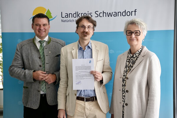 Zwei M&auml;nner und eine Frau stehen vor einem Plakat. Der Mann in der Mitte h&auml;lt eine Urkunde in der Hand.