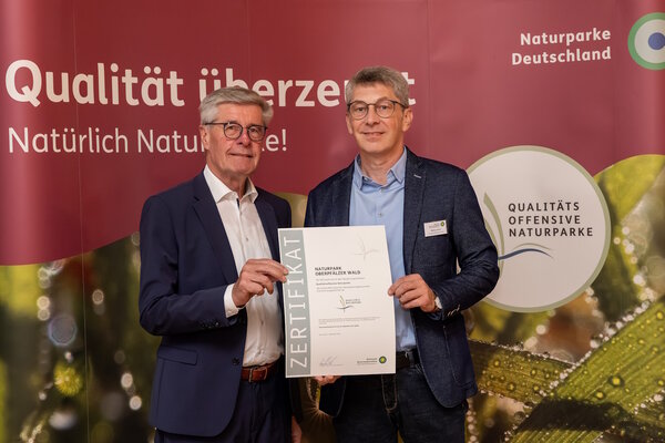 Zwei M&auml;nner stehen vor einem Plakat und halten eine Urkunde in den H&auml;nden