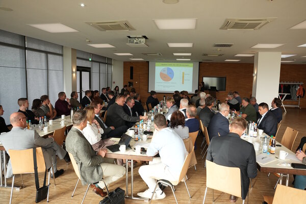 Viele Menschen sitzen an drei langen Tischreihen. An der Wand wird eine Grafik gezeigt.