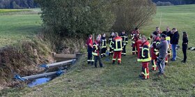 Menschen stehen neben einem Fluss. In dem Fluss liegen &Ouml;lsperren. Viele der Menschen tragen eine Feuerwehr-Ausr&uuml;stung